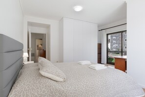 Een slaapkamer met een groot bed, een nachtkastje, een raam met uitzicht op gebouwen en een deur naar een badkamer.
