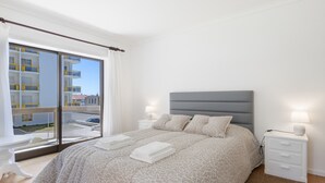 Ein Schlafzimmer mit Bett, Nachttischen, einem Fenster mit Blick auf Gebäude und einem Balkon.