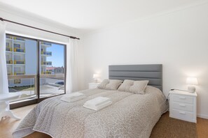 Een slaapkamer met een bed, nachtkastjes, een raam met uitzicht op gebouwen en een balkon.