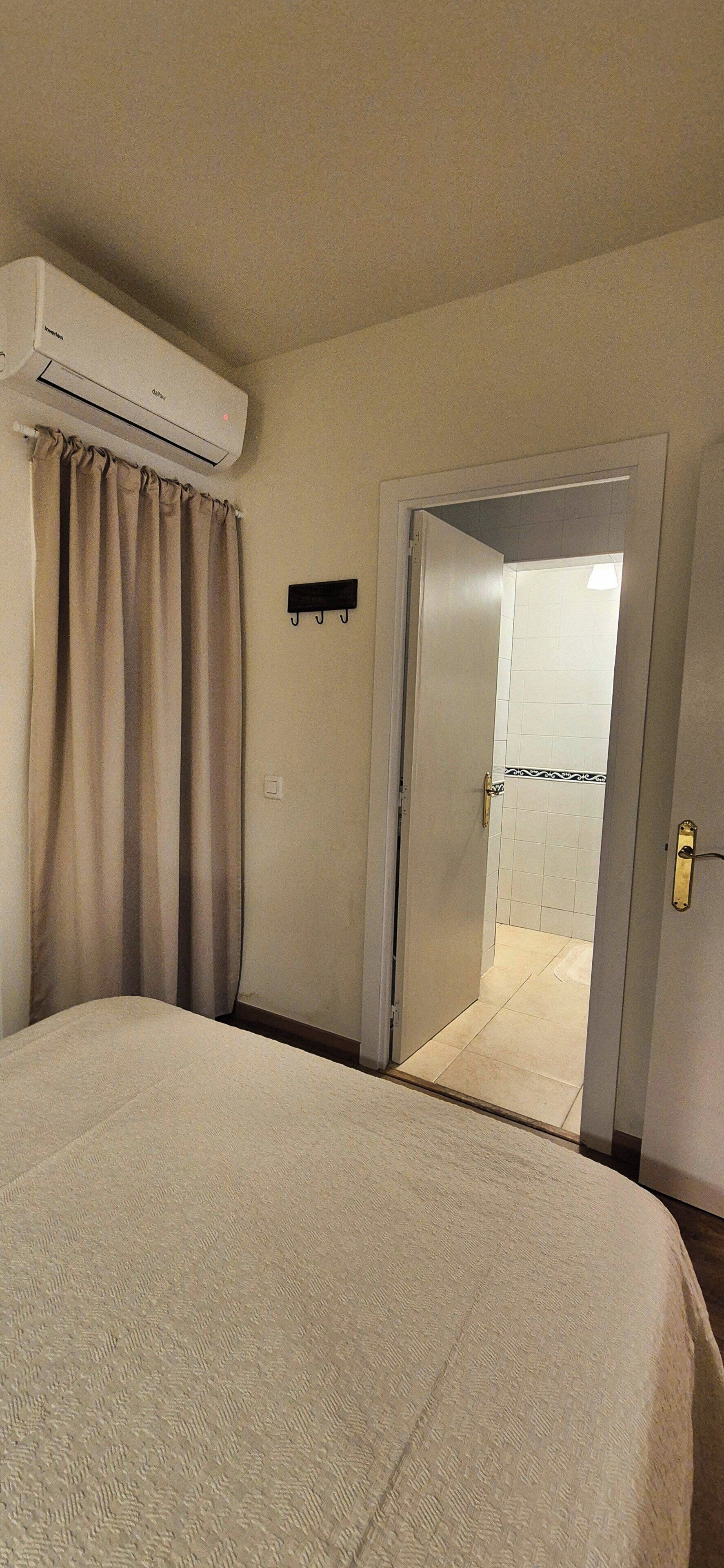 Quarto de hotel com cama, aparelho de ar condicionado, porta para banheiro e porta-toalhas.