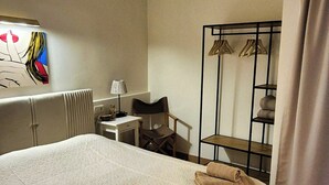 Ein Hotelzimmer mit einem Bett, einem Schreibtisch, einem Stuhl, einem Heizkörper, einem Wandbild und einem Metallgestell mit Kleiderbügeln.