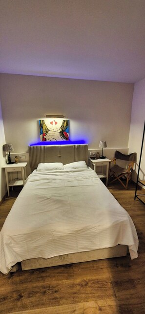 A large bed with a white bedspread, a bedside table with a lamp, a chair, and a wall-mounted artwork.