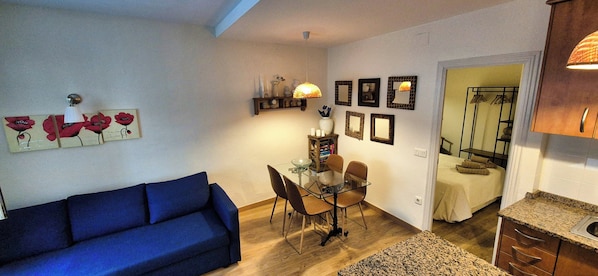 A living room with a blue sofa, a dining area with a glass table and chairs, and a kitchen with granite countertops.