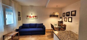 A living room with a blue sofa, a dining table with chairs, a small wooden table, and wall-mounted artwork.