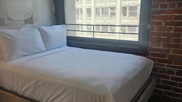 Un letto in una stanza con un'ampia finestra, un muro in mattoni e una parete spoglia con un interruttore della luce.