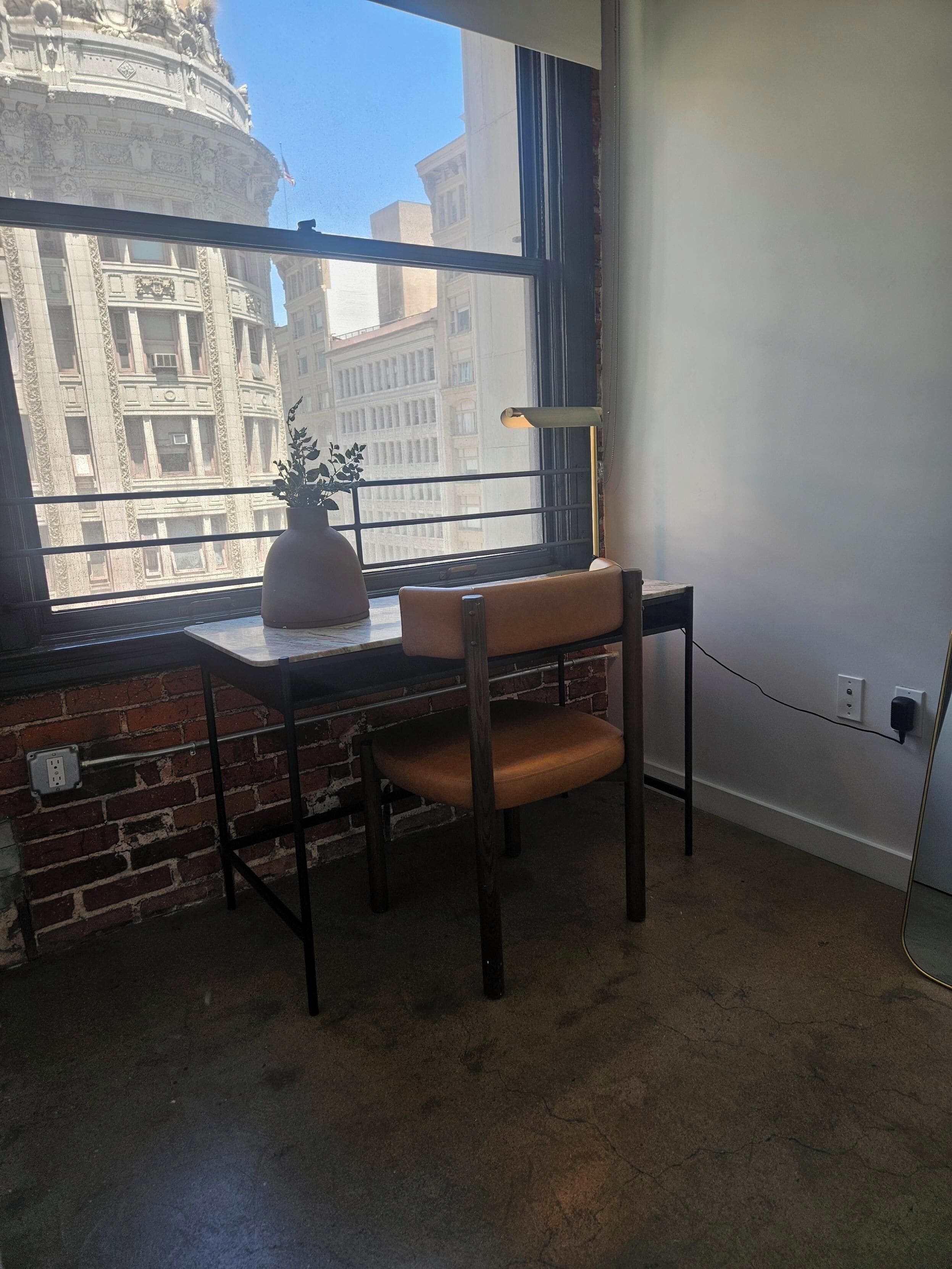 A room with a brick wall, a window with a view of a building, a desk with a chair, and a potted plant.