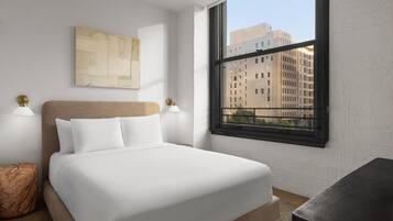 A hotel room with a bed, a window with a view of buildings, and a wall-mounted artwork.