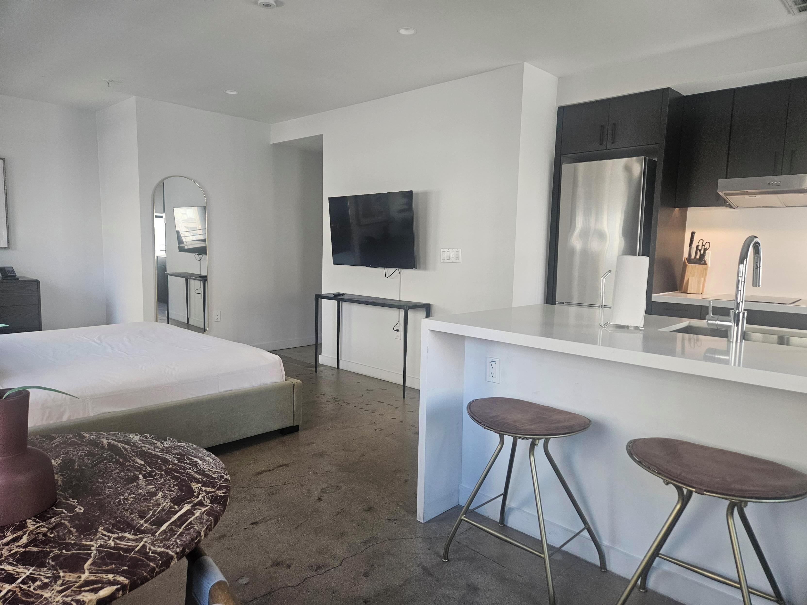 Una habitación de hotel moderna con una cama, televisión, zona de cocina con refrigerador y un taburete pequeño.