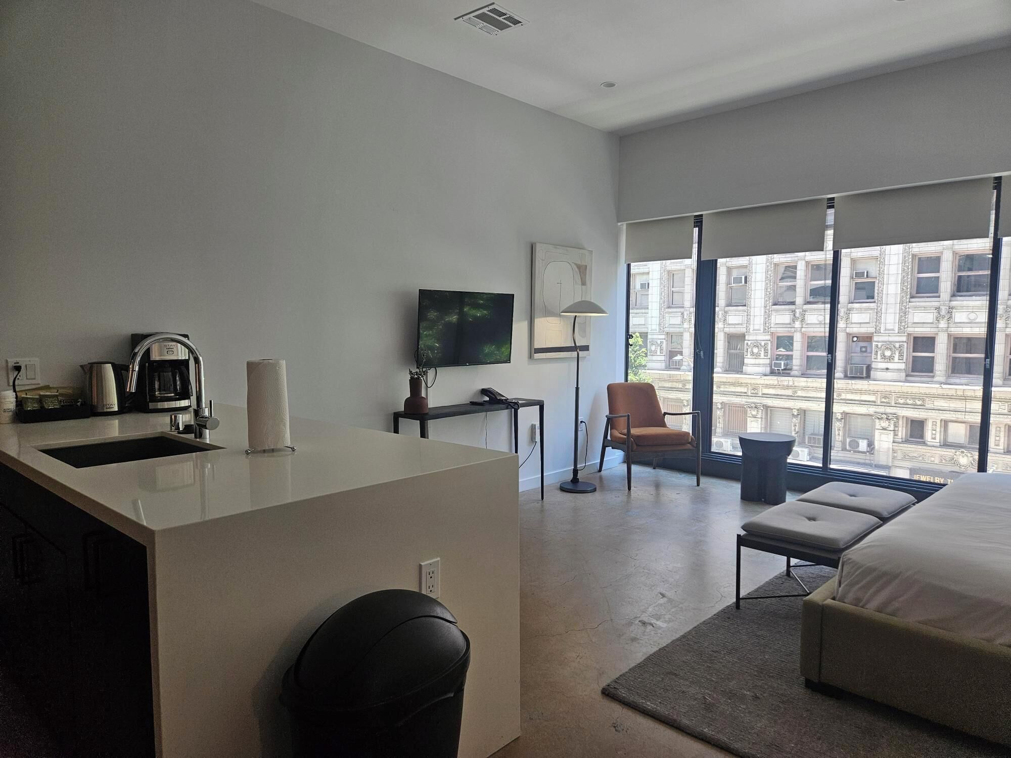 A modern hotel room with a large bed, a desk with a coffee maker, a chair, and a view of buildings through large windows.