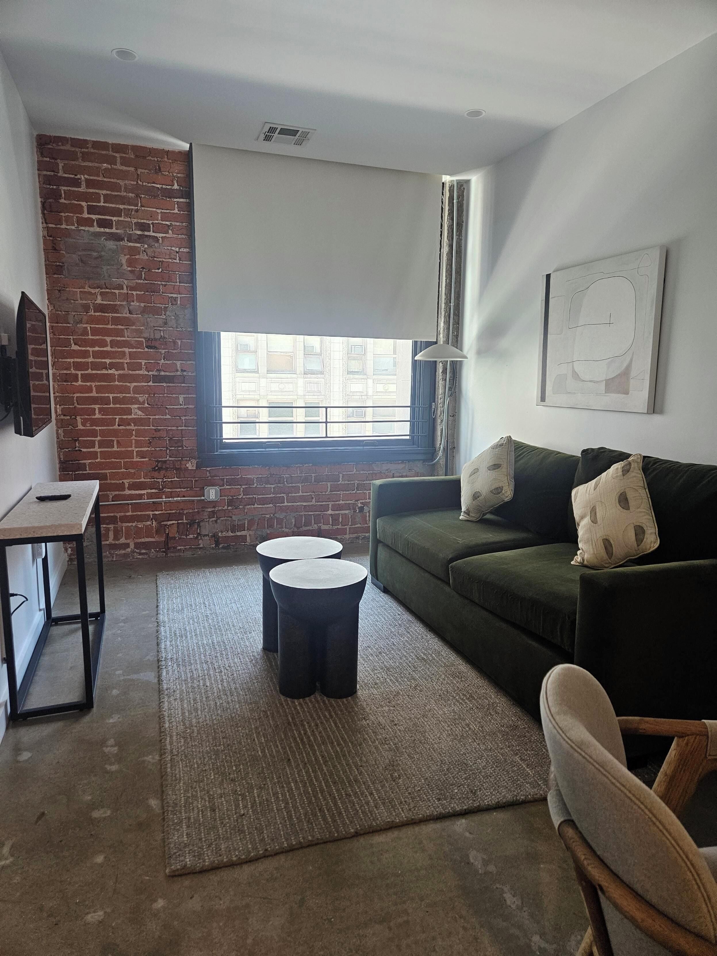 A living room with a brick wall, a green sofa, two round tables, a rug, and a window with blinds.