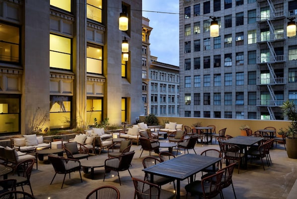 An outdoor patio with seating and tables, surrounded by buildings with large windows and a fire escape.