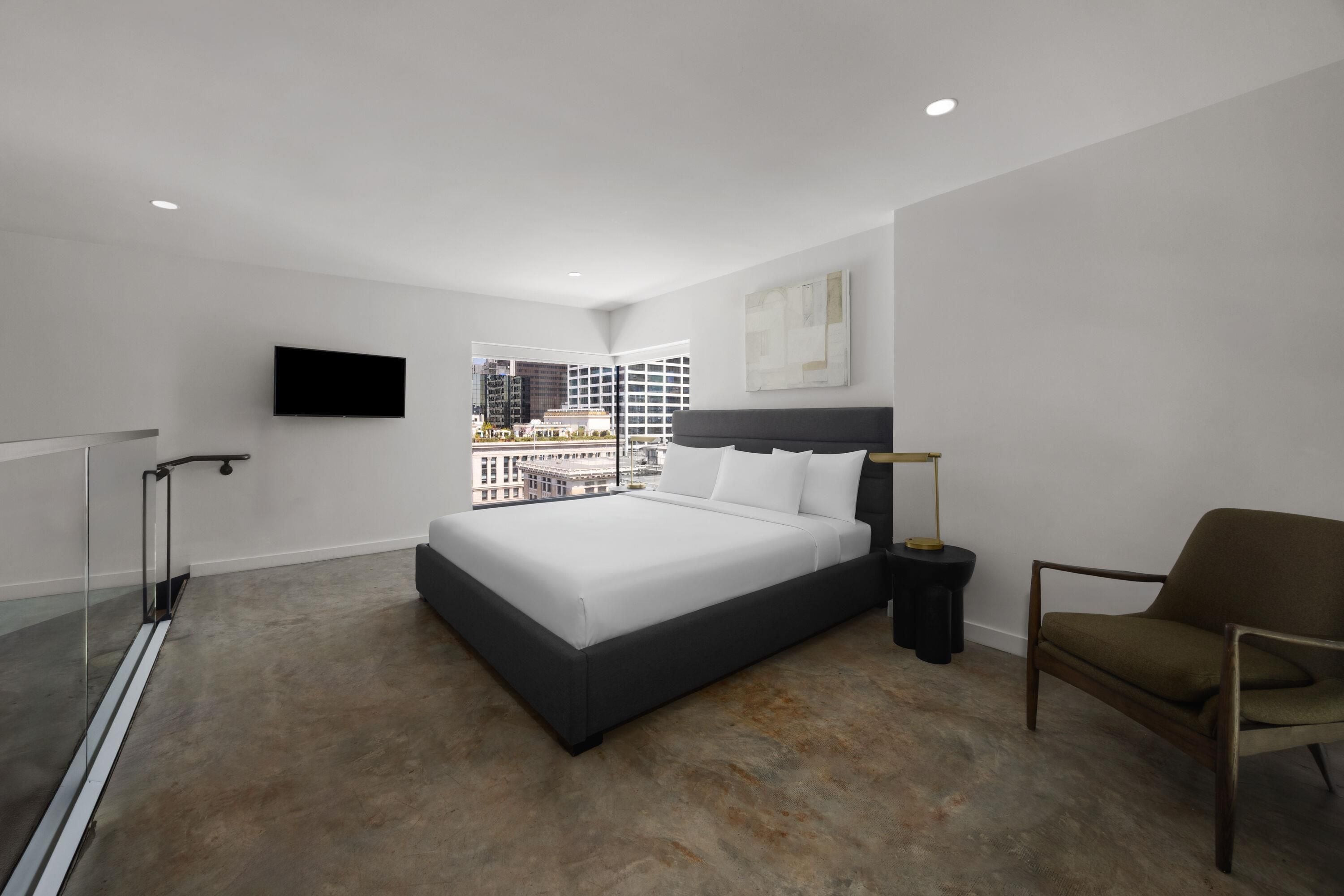 Une chambre d’hôtel moderne dotée d’un grand lit, d’une chaise, d’une table de chevet avec une lampe, d’un téléviseur à écran plat fixé au mur et offrant une vue sur le paysage urbain grâce à de grandes fenêtres.