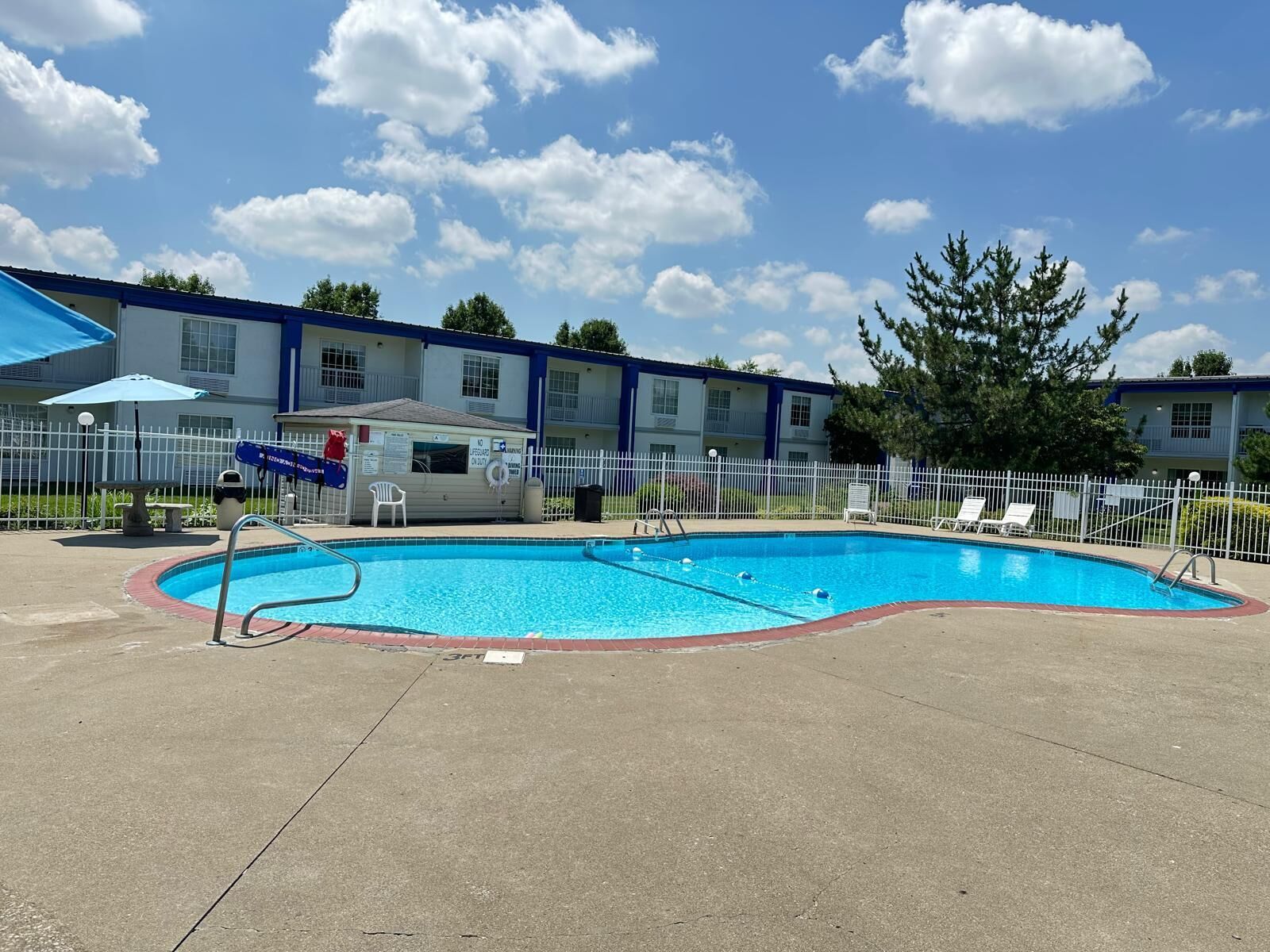 Une piscine entourée d’une clôture blanche, un poste de surveillance pour les sauveteurs et, au fond, un bâtiment aux bordures bleues.