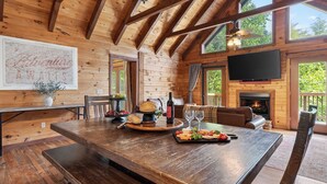 Interno di una baita in legno con un tavolo da pranzo, un camino, una TV e ampie finestre.