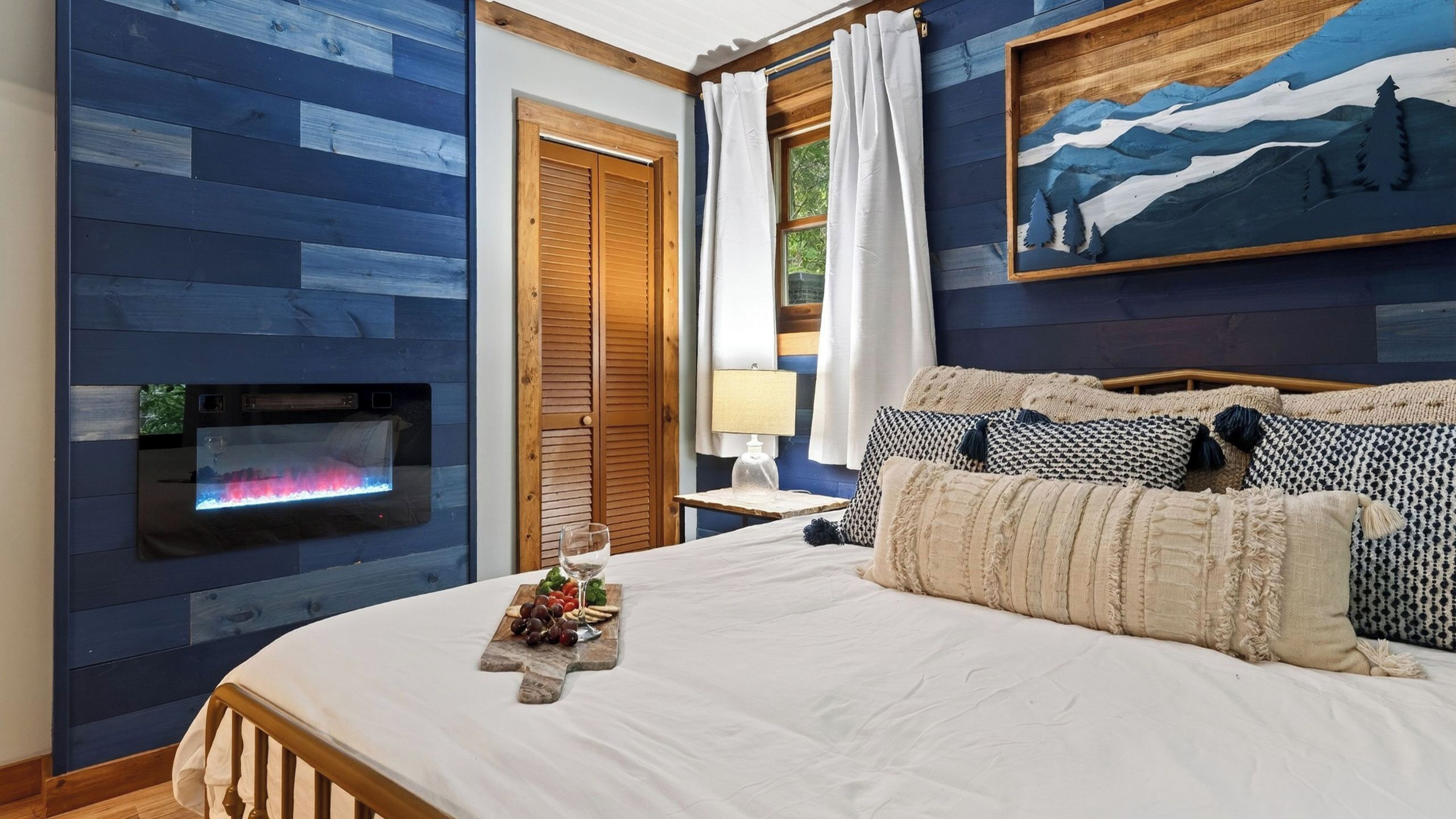 Een slaapkamer met een openhaard, een bed met gestreepte kussens, een nachtkastje met een lamp en een schilderij aan de muur.