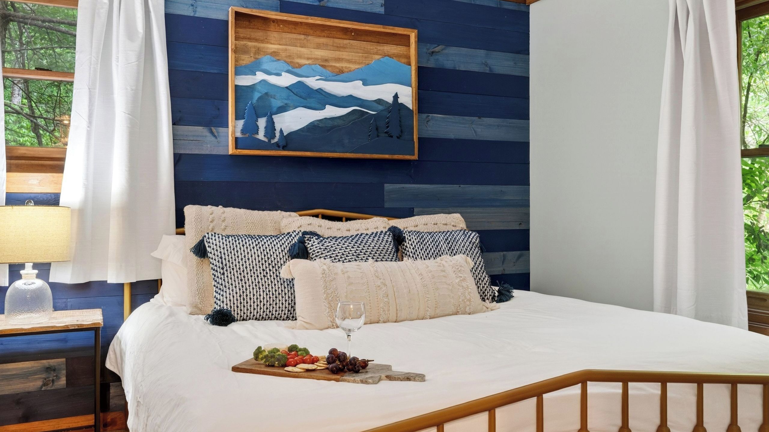 Une chambre à coucher avec un lit, des oreillers, une table de chevet avec une lampe, un tableau et une fenêtre avec des rideaux.