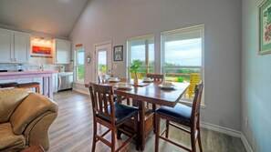 Un comedor con una mesa y sillas de madera, una cocina al fondo y vistas al exterior con vegetación.