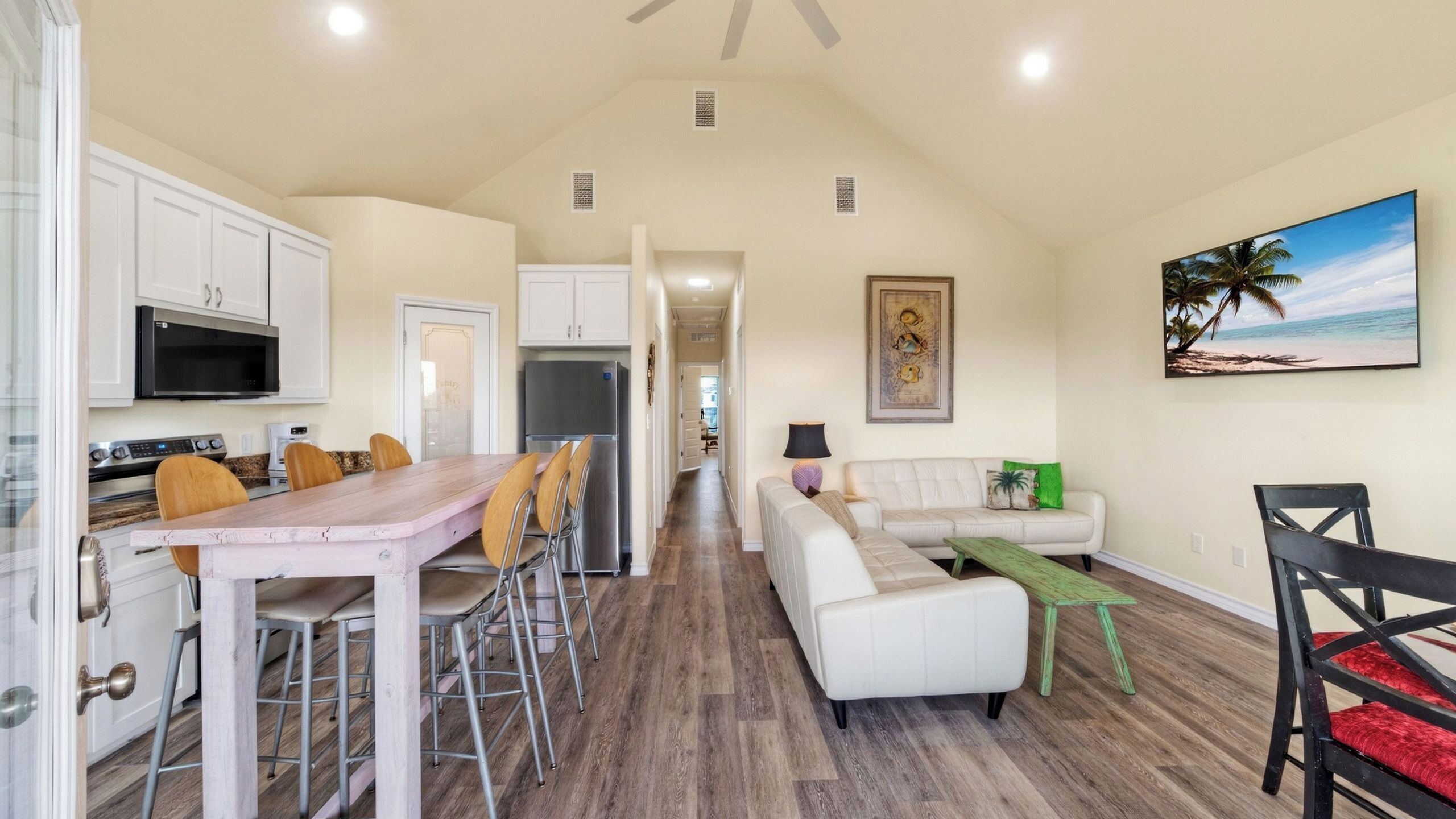 Una cocina amplia con electrodomésticos modernos, un comedor con una mesa y sillas de madera, una sala de estar con sofá y mesa de centro, y un cuadro con motivos playeros en la pared.