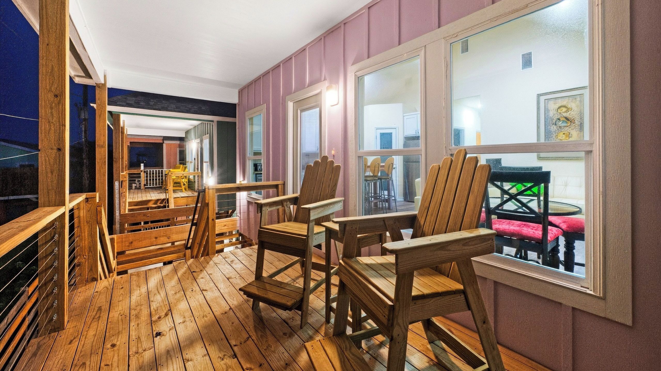 A wooden deck with Adirondack chairs, a dining table, and chairs, and a view of the interior through large windows.