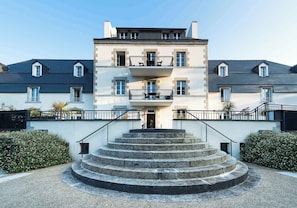 A multi-story building with a central entrance, a staircase leading to the entrance, and a small balcony with outdoor seating.