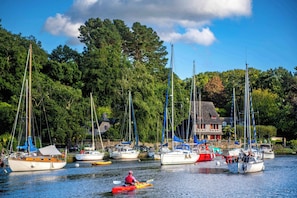 A marina with multiple sailboats docked, a person paddling a kayak, and a building with a sloped roof in the background.