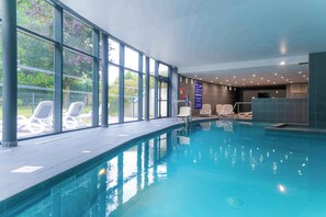 An indoor swimming pool with clear glass walls, lounge chairs, and a reception area.