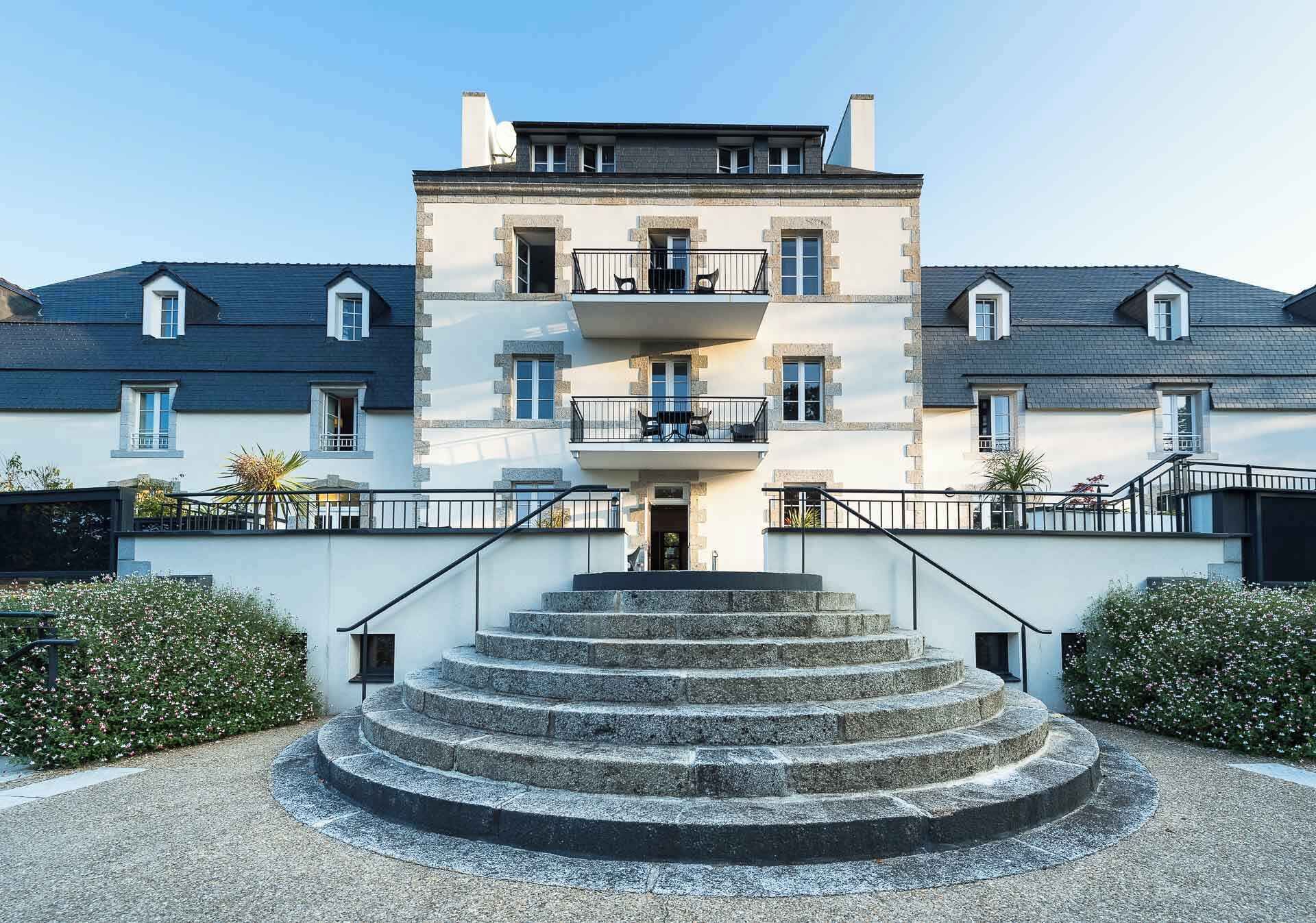Ein mehrstöckiges Gebäude mit einem zentralen Eingang, einer Treppe, die zum Eingang führt, und einem kleinen Balkon im oberen Stockwerk.