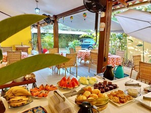Uma área de refeições ao ar livre com móveis de vime, mesas com toalhas xadrez e diversas opções de comida e bebida.