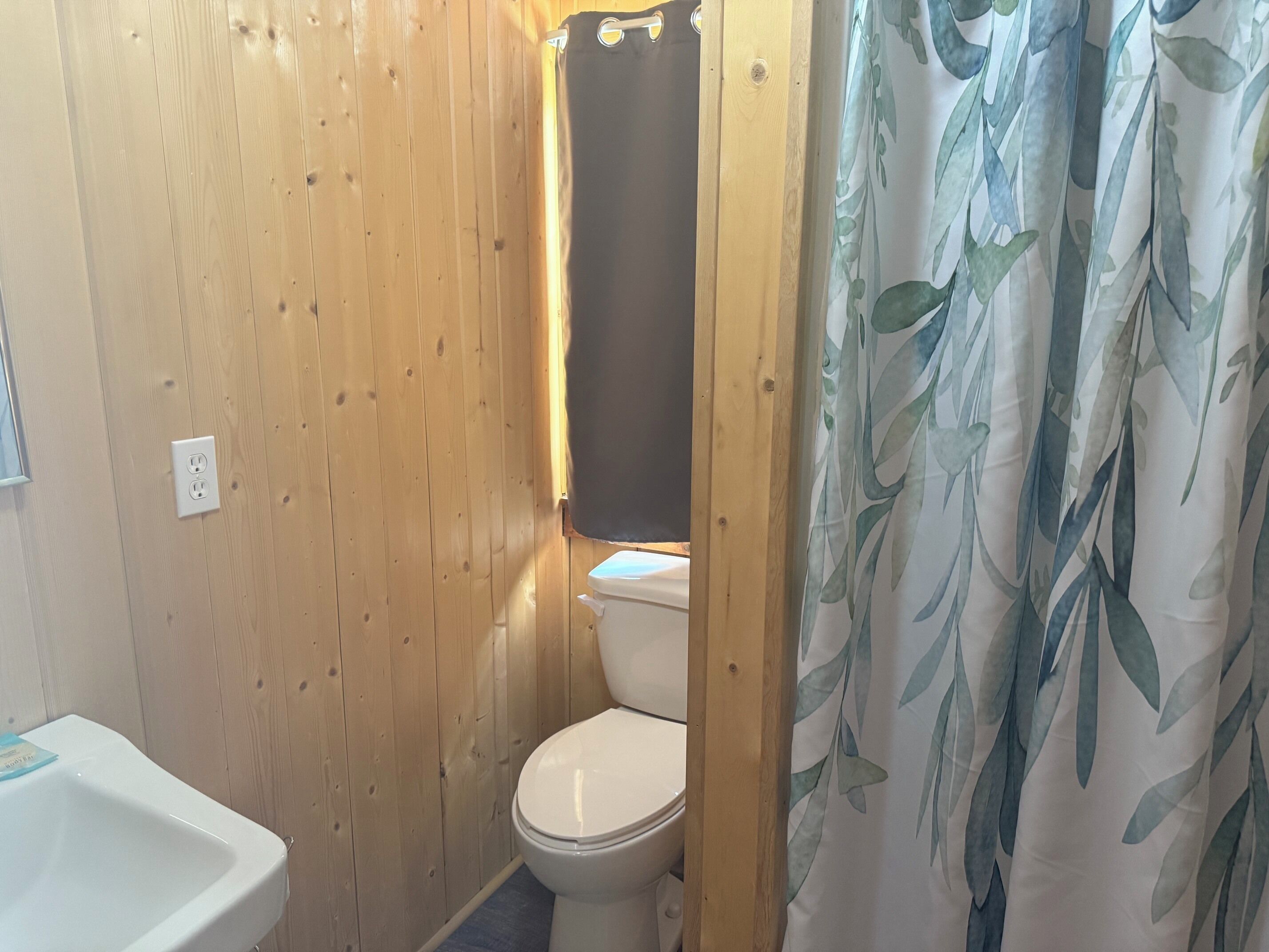 Un baño con paneles de madera, un inodoro blanco, un lavabo blanco y una cortina de ducha con estampado de hojas.