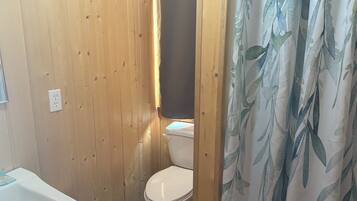 Un baño con paneles de madera, un inodoro blanco, un lavabo blanco y una cortina de ducha con estampado de hojas.
