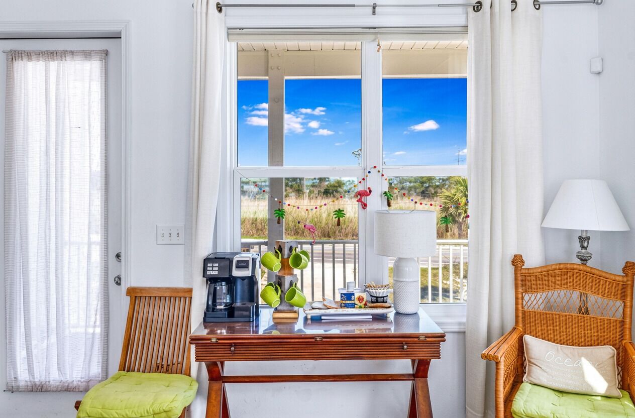 A room with a table, chairs, a coffee maker, a lamp, and a window with a view of a beach and palm trees.