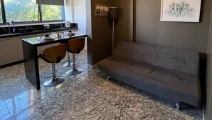 A modern living room with a sofa, a dining table with chairs, a kitchen area, and a large window.