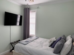 Un dormitorio con cama, televisión de pantalla plana en la pared, ventana con persianas y mesita de noche con lámpara.