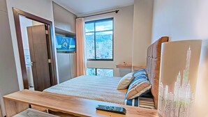 Quarto com cama, televisão na parede, janela com vista para a cidade e mesa de cabeceira de madeira com controle remoto.