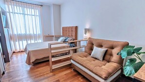 Quarto com cama, sofá, mesa de cabeceira de madeira, janela com cortinas e uma planta em vaso.