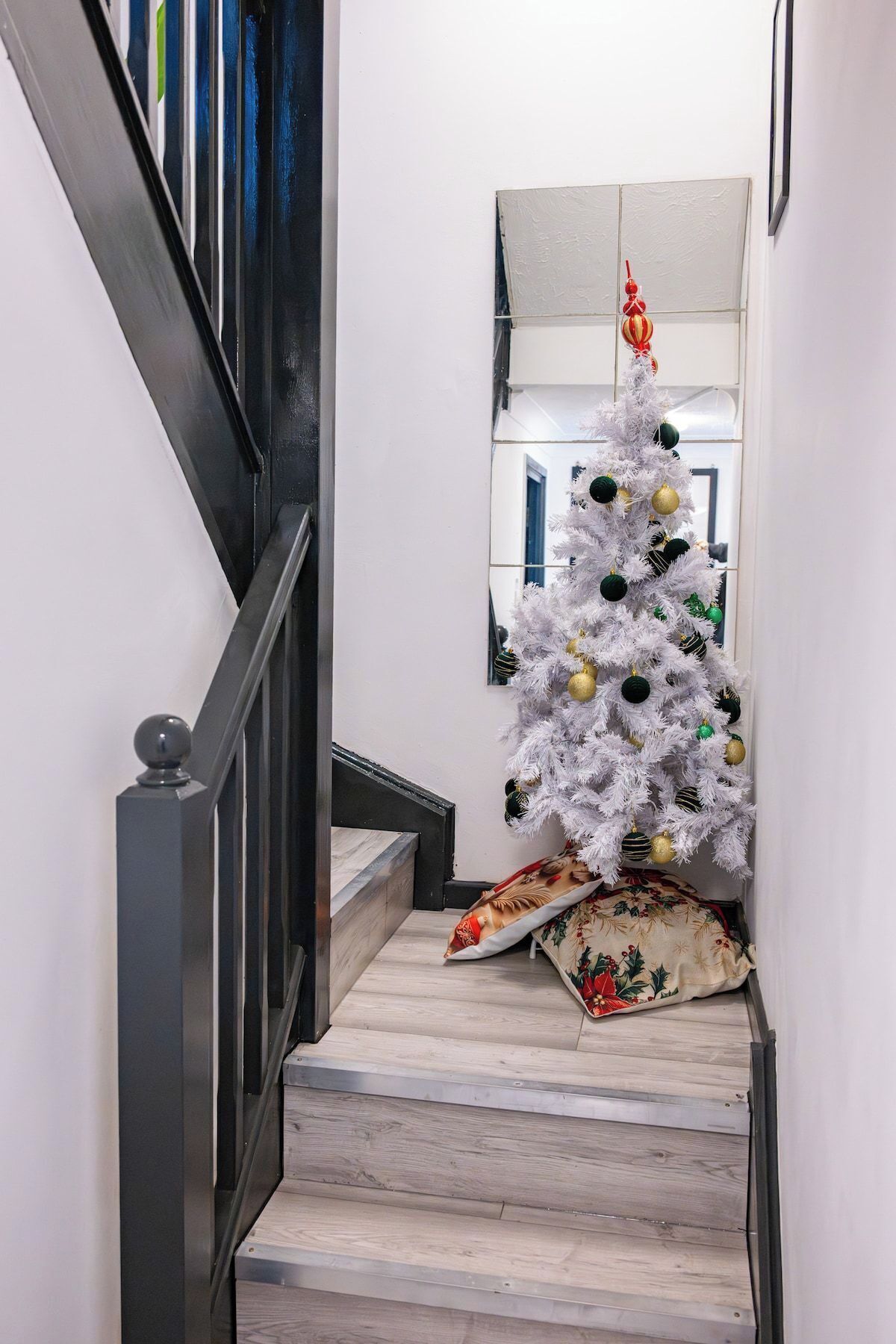 A staircase with a white Christmas tree decorated with gold and green ornaments, leading to a room with a mirror and a framed picture on the wall.