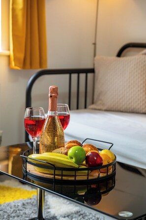 Een bed met hoofdeinde, een nachtkastje met een fles en glazen, en een fruitmand op het tafeltje.