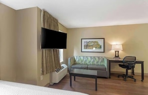A hotel room with a bed, a sofa, a desk with a chair, a TV, a lamp, and a framed picture on the wall.