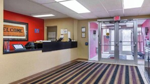 Zona de recepción de hotel con un cartel de 'WELCOME', un mostrador de recepción, una puerta de vidrio con un letrero de 'EXIT' y una alfombra a rayas.