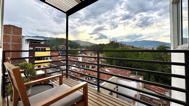 Un balcon avec une chaise en bois et un pot de fleurs, d’où l’on peut admirer un paysage urbain avec des toits en tuiles rouges et des montagnes lointaines.