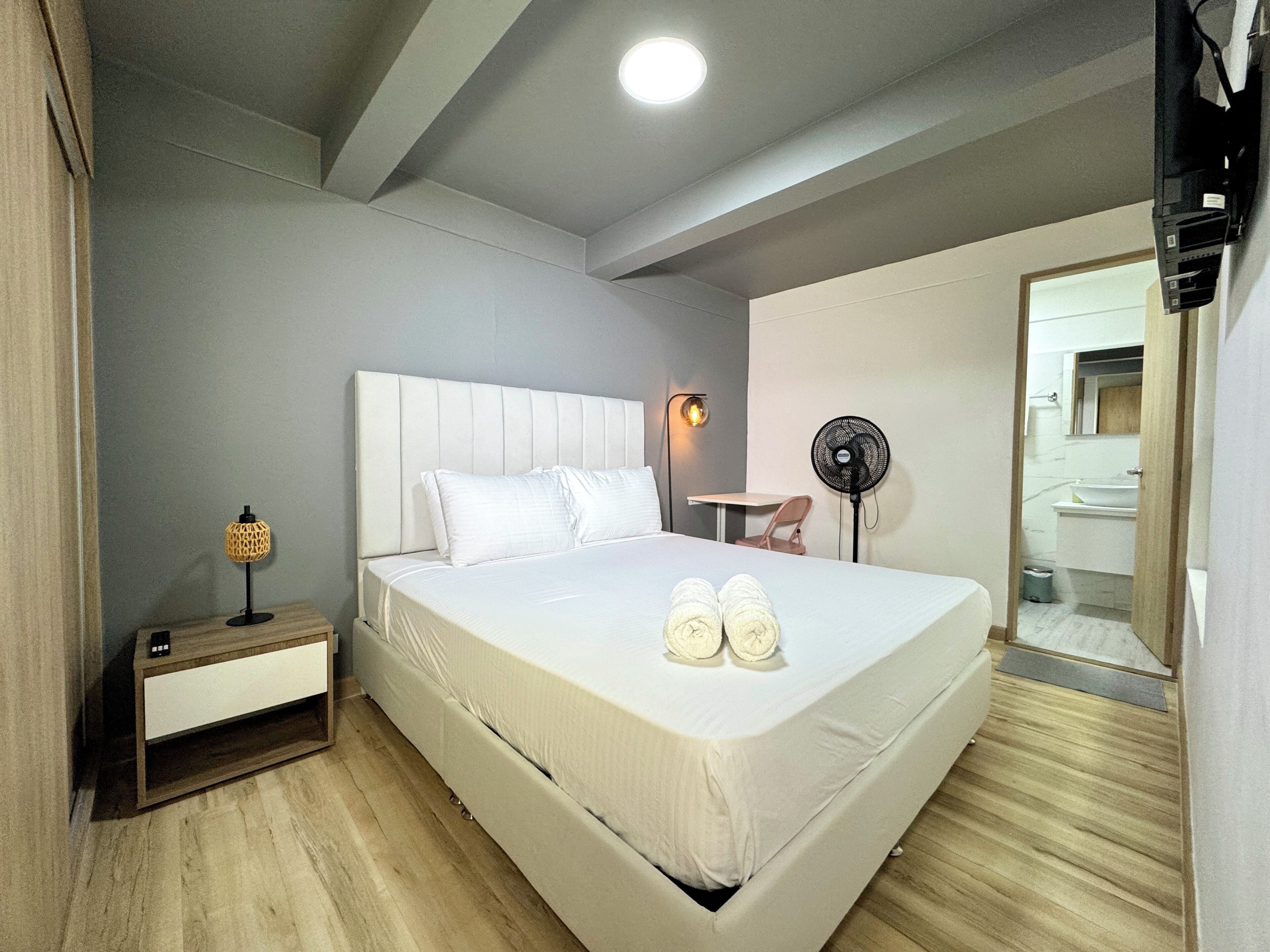 Quarto de hotel com uma cama grande, mesa de cabeceira, luminária e um ventilador.