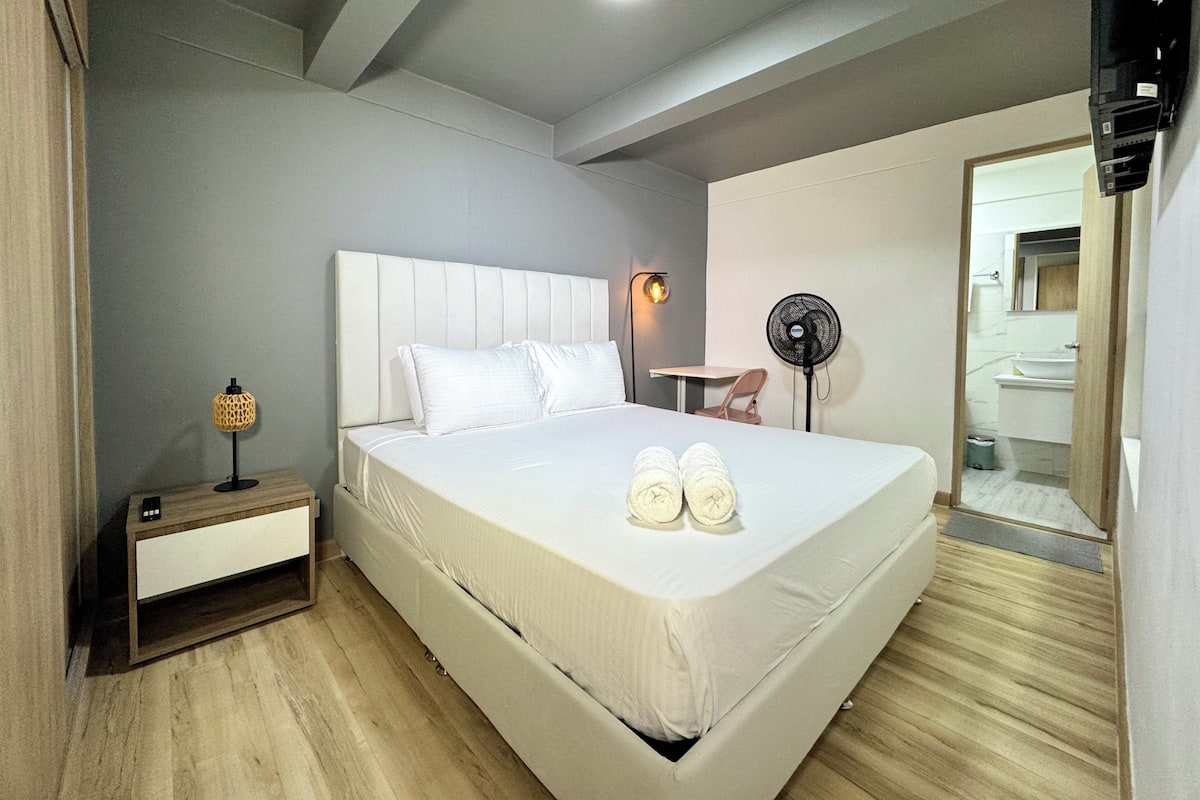 Quarto de hotel com uma cama grande, mesa de cabeceira, luminária e um ventilador.