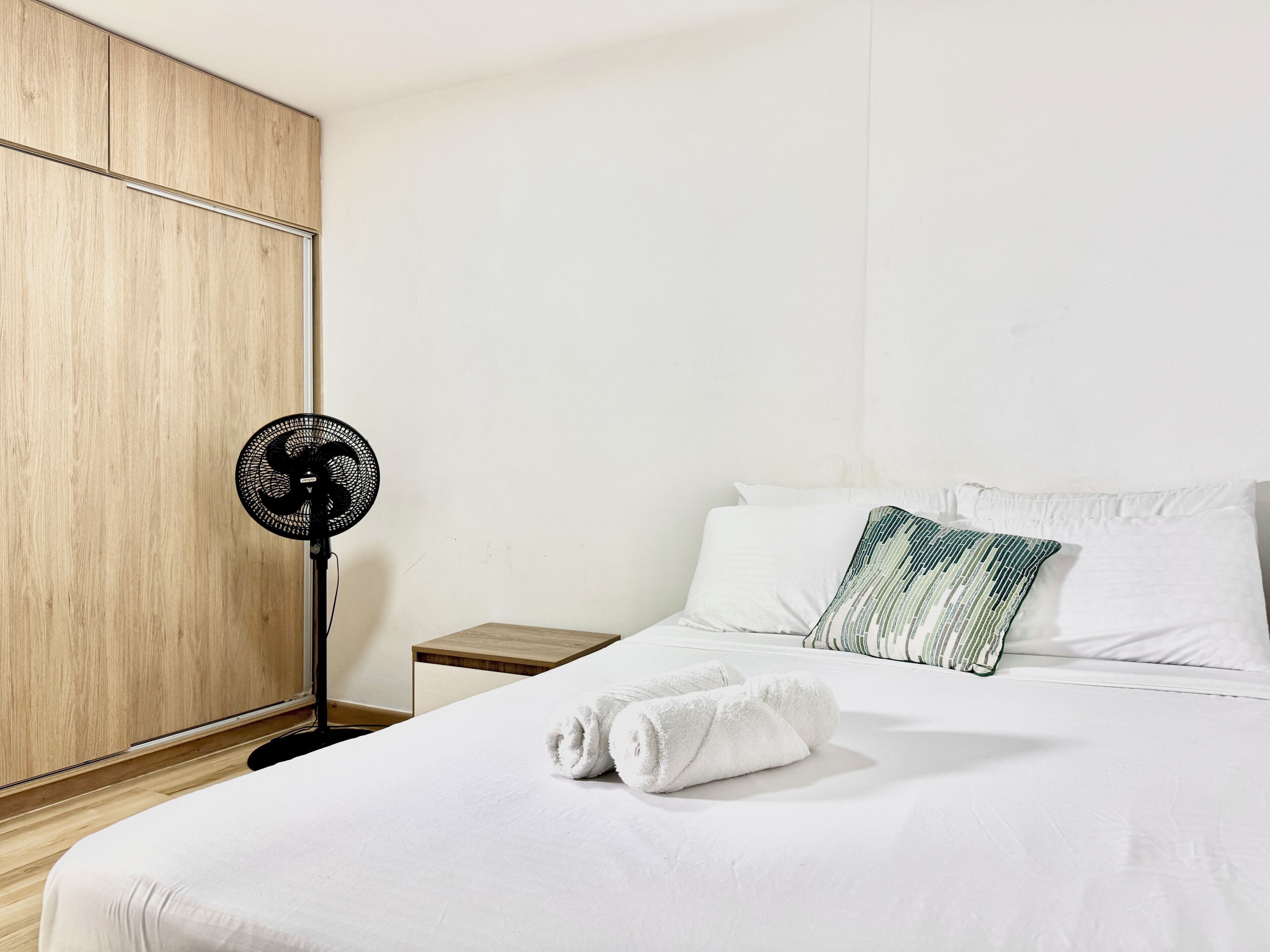 Ein Hotelzimmer mit einem Bett, weißen Bettwäsche, einem Holzschrank, einem Nachttisch, einem Standventilator und einem grün-gestreiften Kissen.