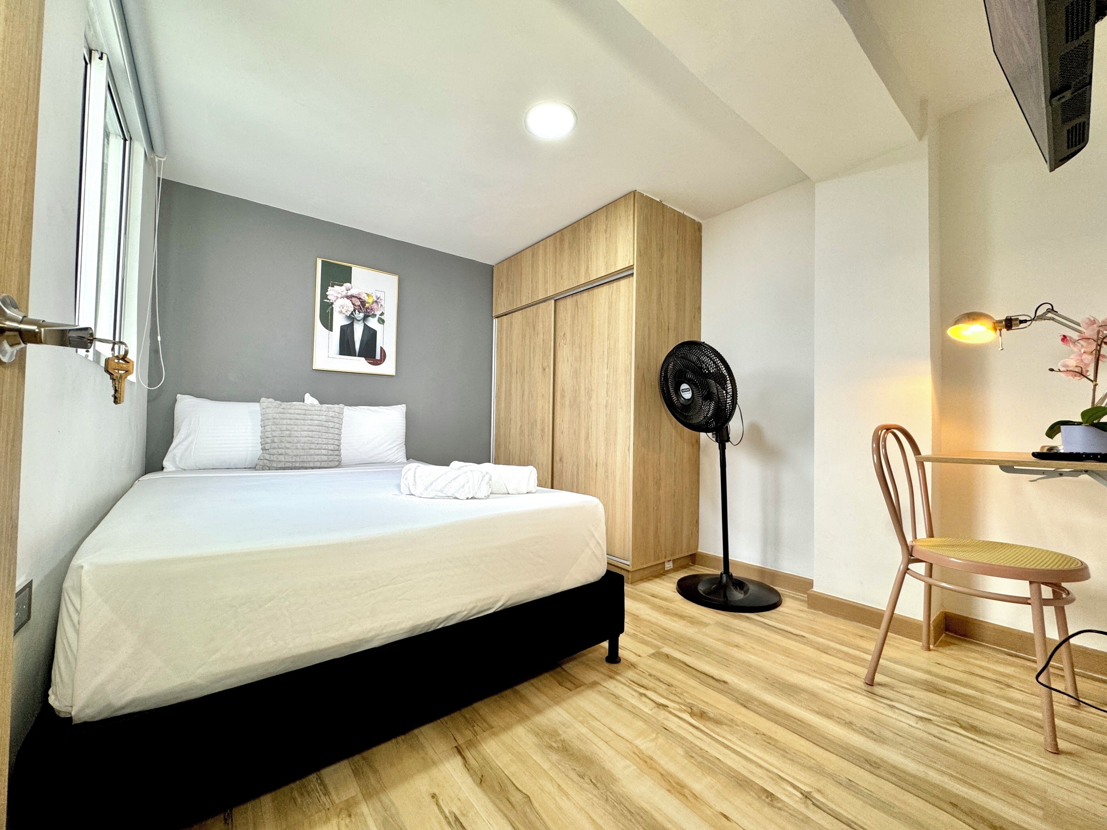 Quarto com uma cama, um ventilador, uma cadeira e uma escrivaninha com abajur.