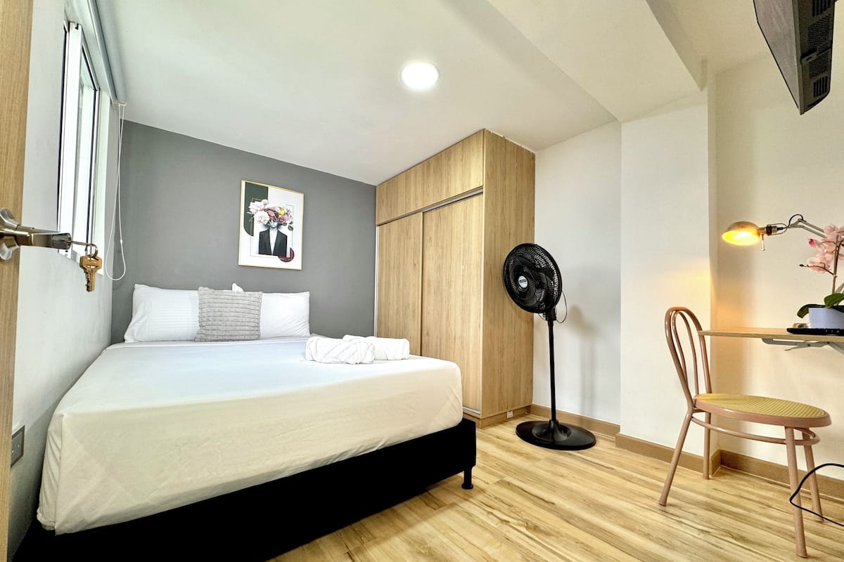 Quarto com uma cama, um ventilador, uma cadeira e uma escrivaninha com abajur.