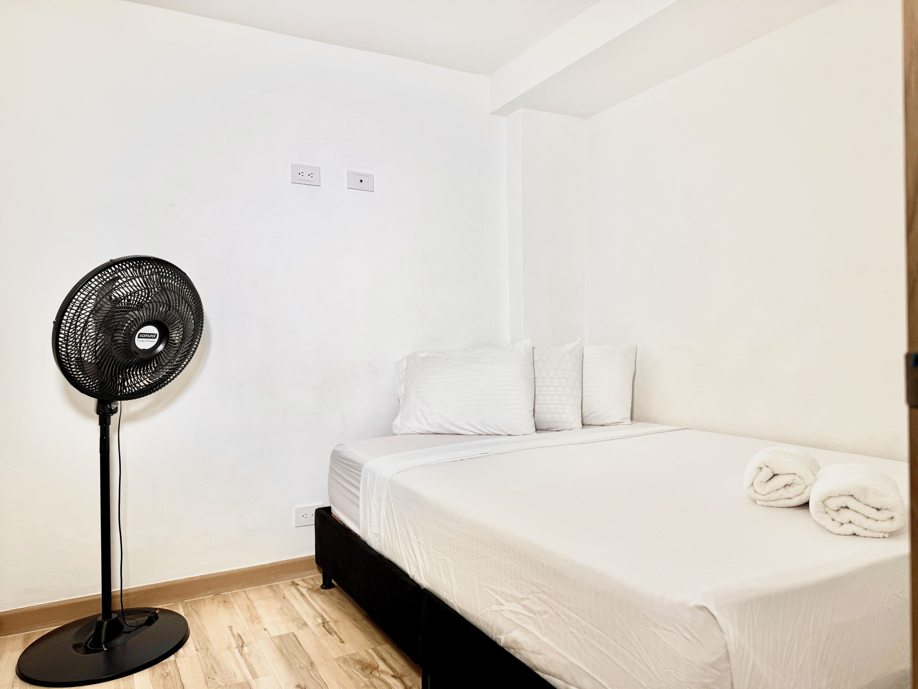 Quarto com cama, roupa de cama branca, ventoinha e piso de madeira.
