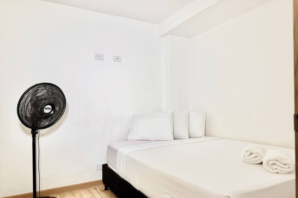 Quarto com cama, roupa de cama branca, ventoinha e piso de madeira.