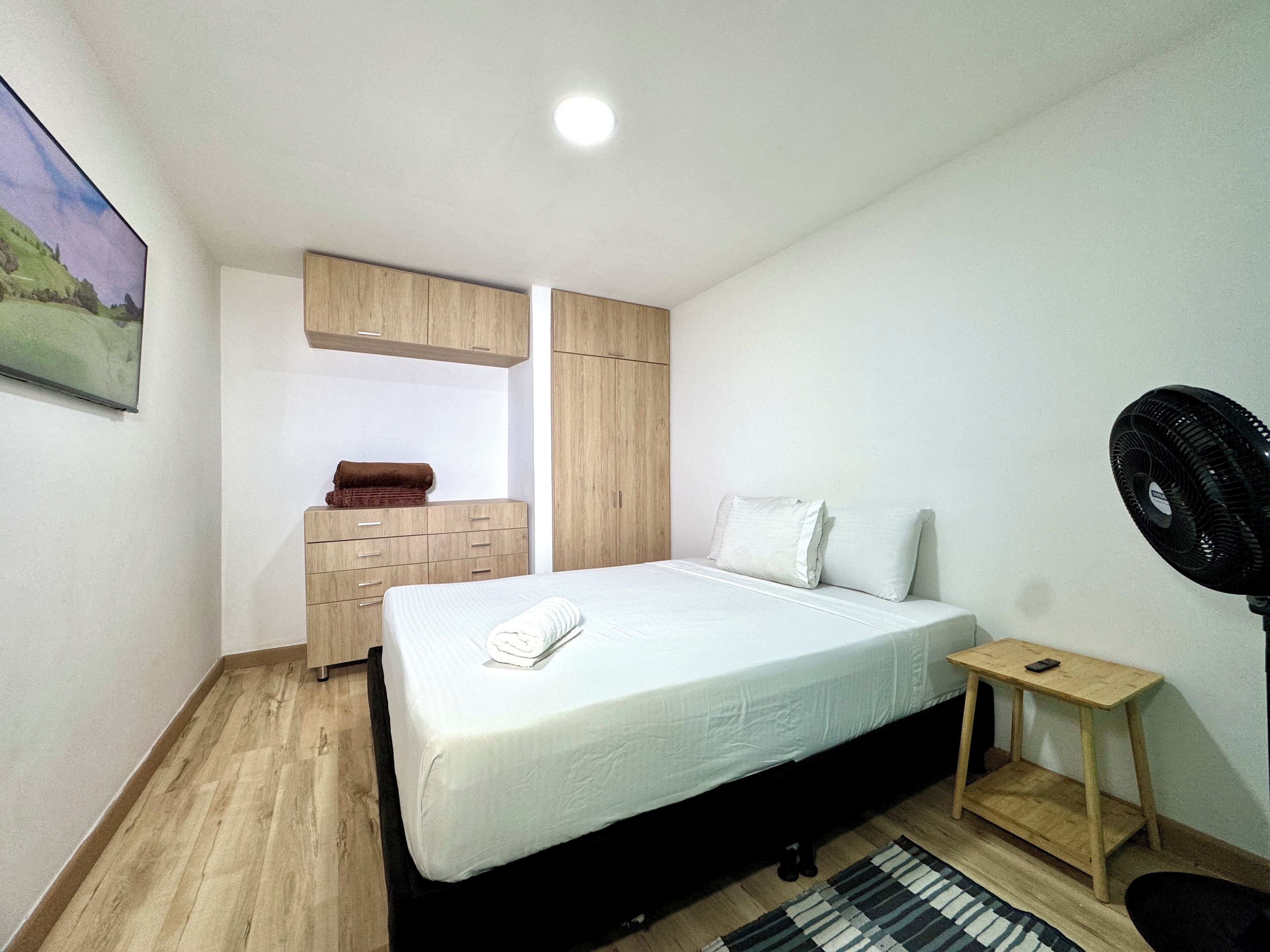 Quarto com cama, guarda-roupa de madeira, uma mesinha com um ventilador e uma janela.