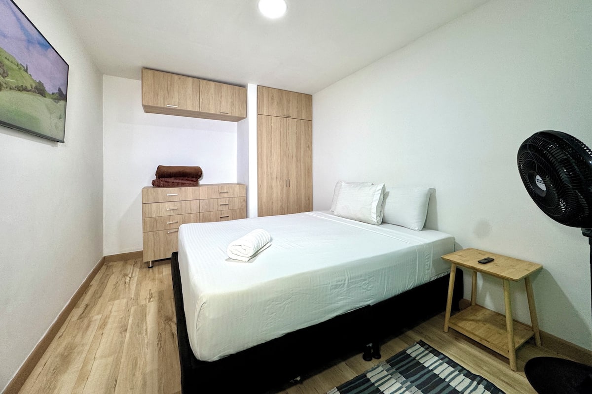Quarto com cama, guarda-roupa de madeira, uma mesinha com um ventilador e uma janela.