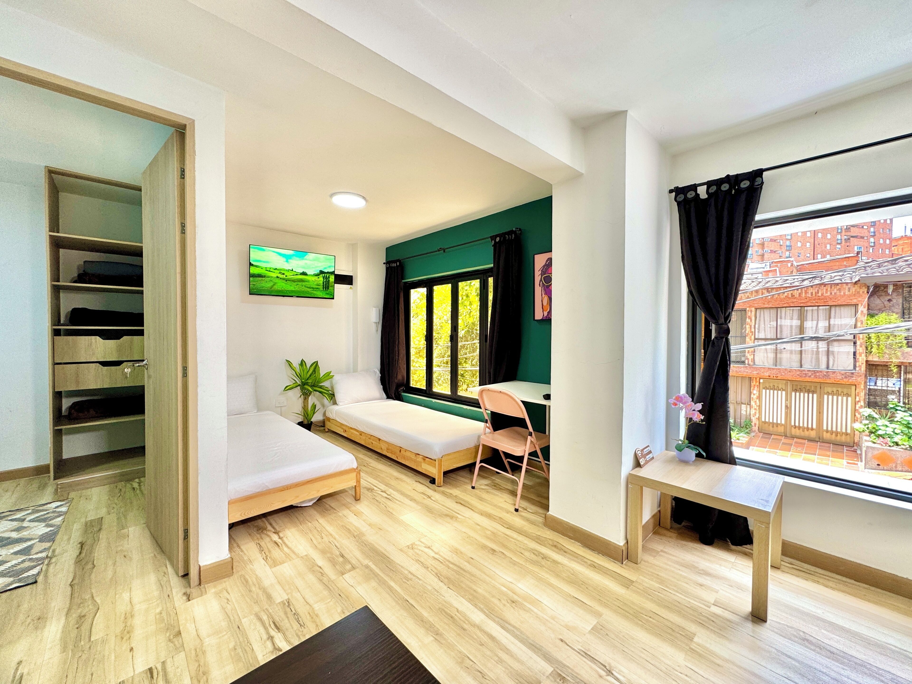 Quarto com cama, escrivaninha, cadeira, televisão e janela com vista para edifícios.